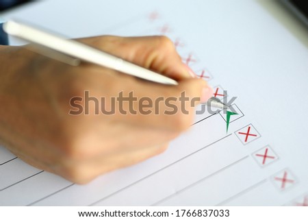 Close-up of person holding silver pen and putting green check on white paper. Application form for personal information. Red crosses and black lines on sheet