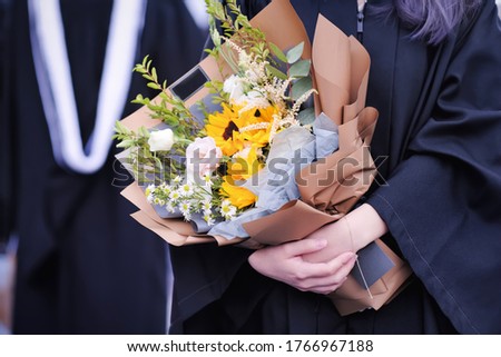 Graduates in academic dresses holding beautiful gorgeous bouquet of fragrant flowers in graduation ceremony at university. Education, new graduation, grad party and people success concept. Close up. Royalty-Free Stock Photo #1766967188
