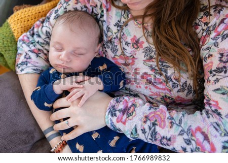 Young mother sits in flowery sweater holds her sleeping infant son. The boy clothed in a blue playsuit rests safely on his mom's chest. 