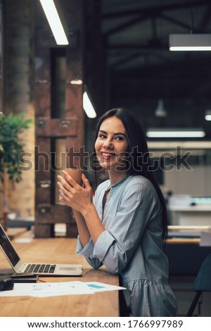 Mirthful female office employee sitting at the table with a laptop and looking away with a smile while holding a mug Royalty-Free Stock Photo #1766997989