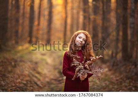 Beautiful red-haired girl with freckles holding a bouquet of dry leaves of fern in her hands. Walk in the autumn forest or park. The atmosphere of early spring or autumn, golden sunset light.
