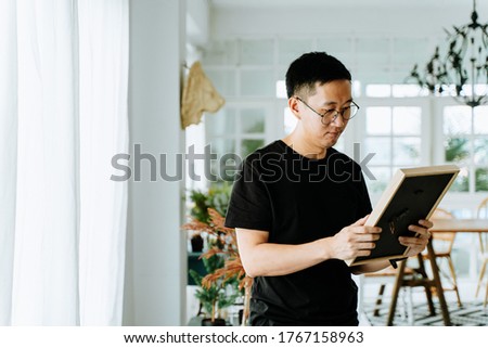 Sad Asian male in glasses looking at framed picture and recollecting memories about loved ones in cozy room at home Royalty-Free Stock Photo #1767158963