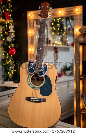 large guitar stands on a wooden table against the background of the Christmas atmosphere. On the neck of the guitar hang a little kid's sneakers. Love for music from birth. New year and Christmas gift