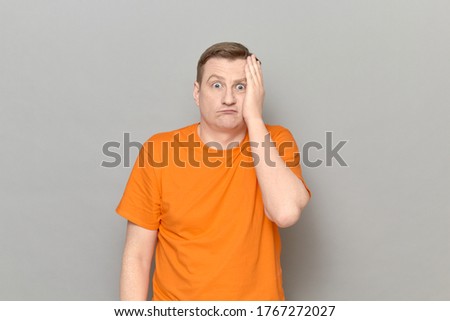 Studio portrait of unhappy confused mature man wearing orange T-shirt, touching his cheek with hand, feeling puzzlement and perplexity, looking like stupid user, standing sideways over gray background