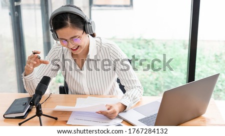 Asian women podcaster podcasting and recording online talk show at studio using headphones, professional microphone and computer laptop on table looking at camera for radio podcast.