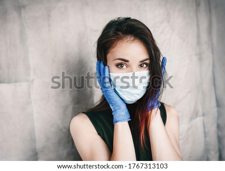 Beautiful terrified young woman in a medical mask and protective surgical sterile gloves on her arm, on gray background, protection against coronovirus.