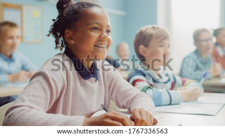 In Elementary School Class: Portrait of a Brilliant Black Girl with Braces Writes in Exercise Notebook, Smiles. Junior Classroom with Diverse Group of Children Learning New Stuff Royalty-Free Stock Photo #1767336563