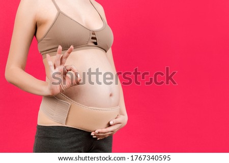 Close up of pregnant woman wearing supporting bandage and showing okay gesture at pink background with copy space. Orthopedic abdominal support belt concept.