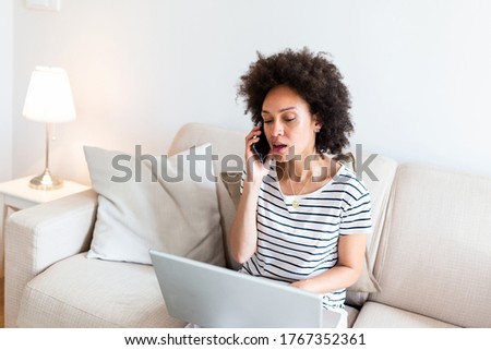 woman sitting on sofa with laptop and talking on phone at home. Young successful businesswoman working from home while talking at phone. College student studying on laptop and using phone.