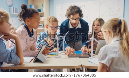 Elementary School Classroom: Enthusiastic Teacher Holding Tablet Computer Explains Lesson to Brilliant Young Children. Kids Learning Programming Languages, Internet Safety and Software Design Royalty-Free Stock Photo #1767364385