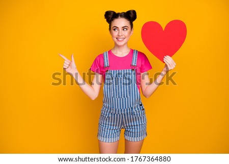 Photo of pretty funny lady hold big paper heart showing creative date invitation direct finger look empty space wear magenta t-shirt denim overall isolated yellow bright color background