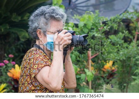 A portrait of an elderly woman wearing a face mask and shooting photo by digital camera while standing in a garden. New normal. Concept of old people, healthcare and photography