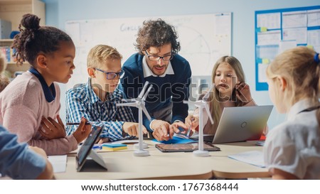 Elementary School Classroom: Enthusiastic Teacher Holding Tablet Computer Explains to a Brilliant Young Children How Wind Turbines Work. Kids Learning about Eco-Friendly Forms of Renewable Energy Royalty-Free Stock Photo #1767368444