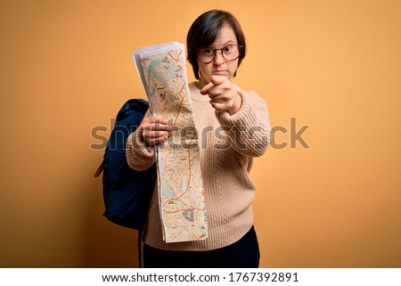 Young down syndrome tourist woman wearing bag and looking at destination city map pointing with finger to the camera and to you, hand sign, positive and confident gesture from the front