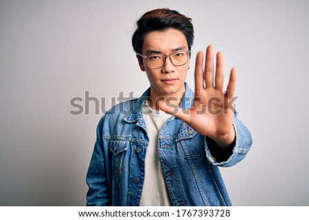 Young handsome chinese man wearing denim jacket and glasses over white background doing stop sing with palm of the hand. Warning expression with negative and serious gesture on the face.