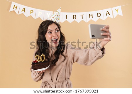Image of excited woman taking selfie on cellphone with birthday cake isolated over beige background