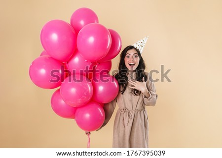 Image of excited cute woman in party cone smiling while posing with pink balloons isolated over beige background