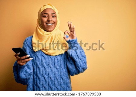Young African American afro woman wearing muslim hijab using smartphone doing ok sign with fingers, excellent symbol