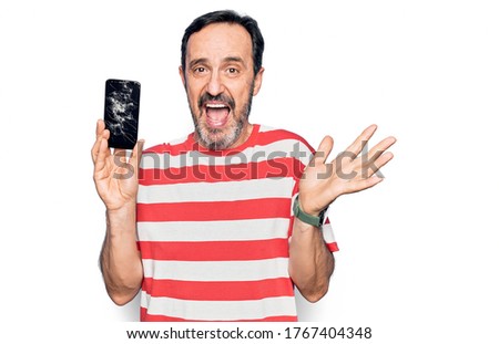 Middle age man holding broken smartphone showing cracked screen over white background celebrating achievement with happy smile and winner expression with raised hand