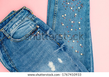 Denim jeans pants with beads on pink surface. Ripped Destroyed Torn Blue denim cloth background.  Royalty-Free Stock Photo #1767410933