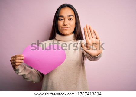 Young beautiful woman holding pink heart standing over isolated background with open hand doing stop sign with serious and confident expression, defense gesture