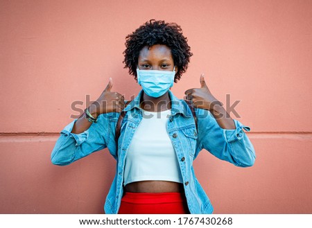 nice black girl, smiling with the eyes and wearing protective and medical mask, making the gesture of thumbs up, positive and optimism, over pastel coral pink background, vignetting effect