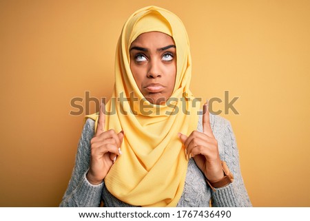 Young beautiful african american girl wearing muslim hijab over isolated yellow background Pointing up looking sad and upset, indicating direction with fingers, unhappy and depressed.