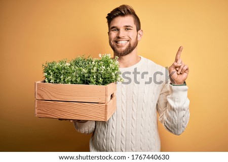 Young blond gardener man with beard and blue eyes holding wooden box with plants surprised with an idea or question pointing finger with happy face, number one