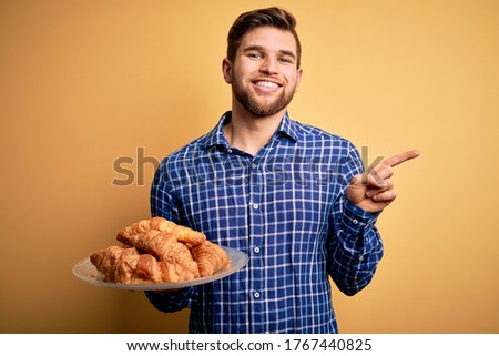 Young blond man with beard and blue eyes holding plate with french croissants to breakfast very happy pointing with hand and finger to the side