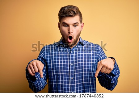 Young blond businessman with beard and blue eyes wearing shirt over yellow background Pointing down with fingers showing advertisement, surprised face and open mouth