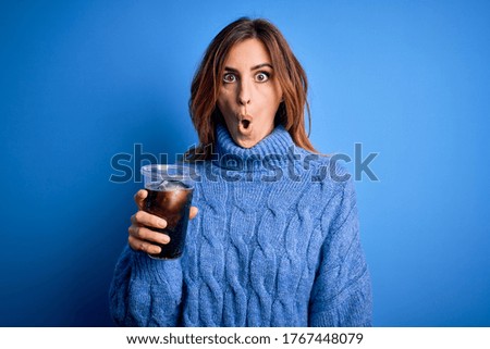Young beautiful brunette woman drinking glass with cola refreshment over blue background scared in shock with a surprise face, afraid and excited with fear expression