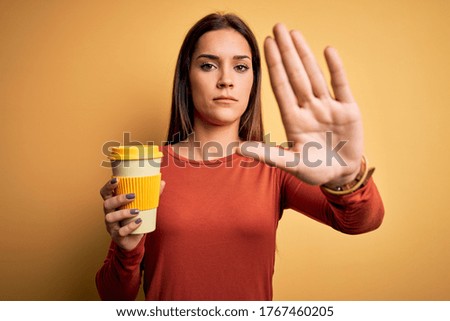 Young beautiful brunette woman drinking cup of takeaway coffe over yellow background with open hand doing stop sign with serious and confident expression, defense gesture