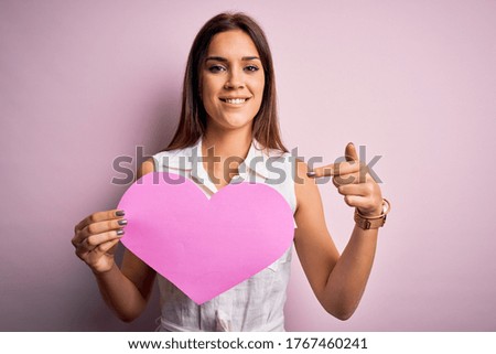 Young beautiful brunette woman holding big pink heart paper celebrating valentine day very happy pointing with hand and finger