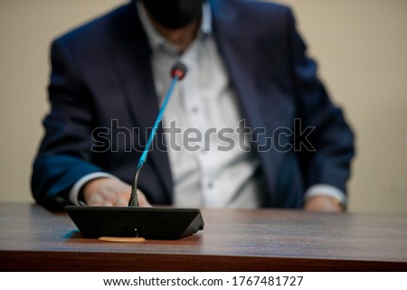 A man in a suit speaks while sitting at a table during a business conference or negotiations with business partners. Included microphone and report. Work time. Business style. Shallow depth of field.