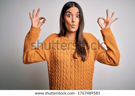 Young beautiful brunette woman wearing casual sweater over isolated white background looking surprised and shocked doing ok approval symbol with fingers. Crazy expression