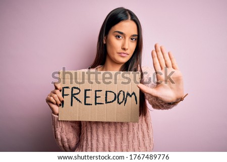 Young beautiful brunette activist woman protesting for freedom holding poster with open hand doing stop sign with serious and confident expression, defense gesture