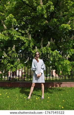 Street fashion concept: full body portrait of young beautiful woman standing in the green park. Model looking aside. Caucasian lady wearing grey silver dress coat, shiny trendy shoes. Fashion concept