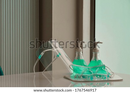 Transparent plastic faceshields, green sanitizers and green medical mask on white table in front of the glass door for wearing on face and rubbing hands, concept for anticoronavirus or covid-19.
