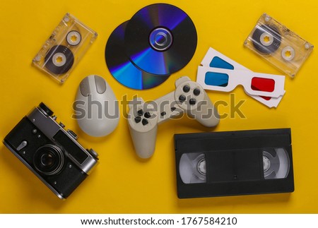 Retro entertainment. Attributes, gadgets 80s. Keyboard, pc mouse, compact discs, gamepad, anaglyph glasses, audio and video cassette, camera on yellow background. Top view. Flat lay