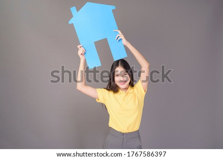 portrait of young confident beautiful woman wearing yellow shirt is holding a blue paper house on grey background studio