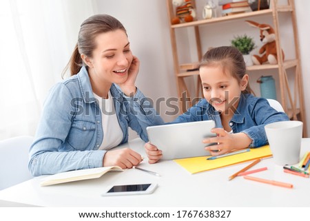 Family concept. Mother and daughter play on digital tablet, spent time together at home, doing homework, read new kids book. Little girl smile have fun with tutor sister. Two person sit at table. Cute