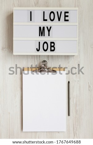 'I love my job' on a lightbox, clipboard with blank sheet of paper on a white wooden background, top view. Flat lay, from above, overhead. 