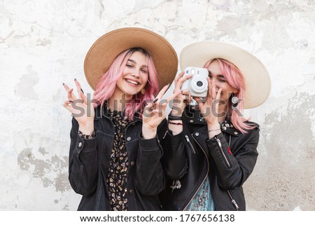 Pretty funny girl with pink hair with smile in straw hat in fashion jacket stands next to sister with modern camera in jacket and shows sign of peace. Lovely happy girls takes positive snapshots.