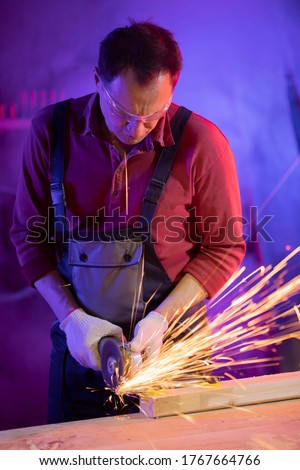 handsome middle-aged male worker in jumpsuit protective goggles and gloves grinding metal with sparks in coloured light in garage metalwork concept jack of all trades concept