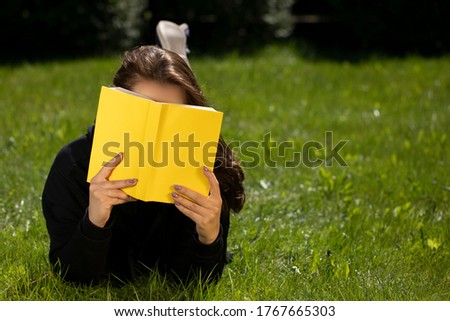 Attractive brunette girl with long hair dressed in black hoodie lying on grass of green lawn reading yellow book on nice summer day. Relaxed vacation concept copy space. Distance Education Concept