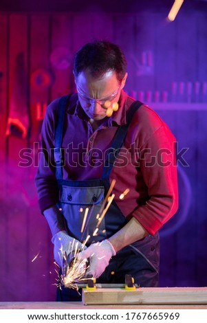 handsome middle-aged male craftsman in jumpsuit protective goggles and gloves grinding metal with sparks in coloured light in garage metalwork concept jack of all trades concept