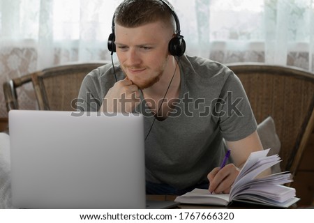 attractive thinking young man in headphones works with laptop, communicates in social networks, makes notes in diary in country wooden house on background of white curtain online conference concept