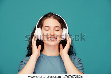 Smiling young woman with closed eyes holding headphones while enjoying favorite song against blue background