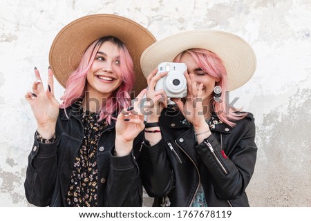 Two fashion charming positive girlfriends with pink hair in trendy youth clothes in straw stylish hats smiling and taking pictures with modern camera. Positive funny girls photographers outdoors.