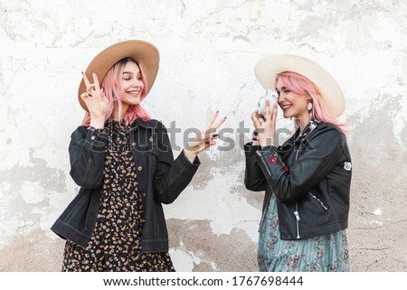 Modern young woman photographer takes snapshot positive girl with cute smile with pink hair in straw hat in denim jacket that shows peace sign. Fashion girlfriends take photo on vintage camera in city